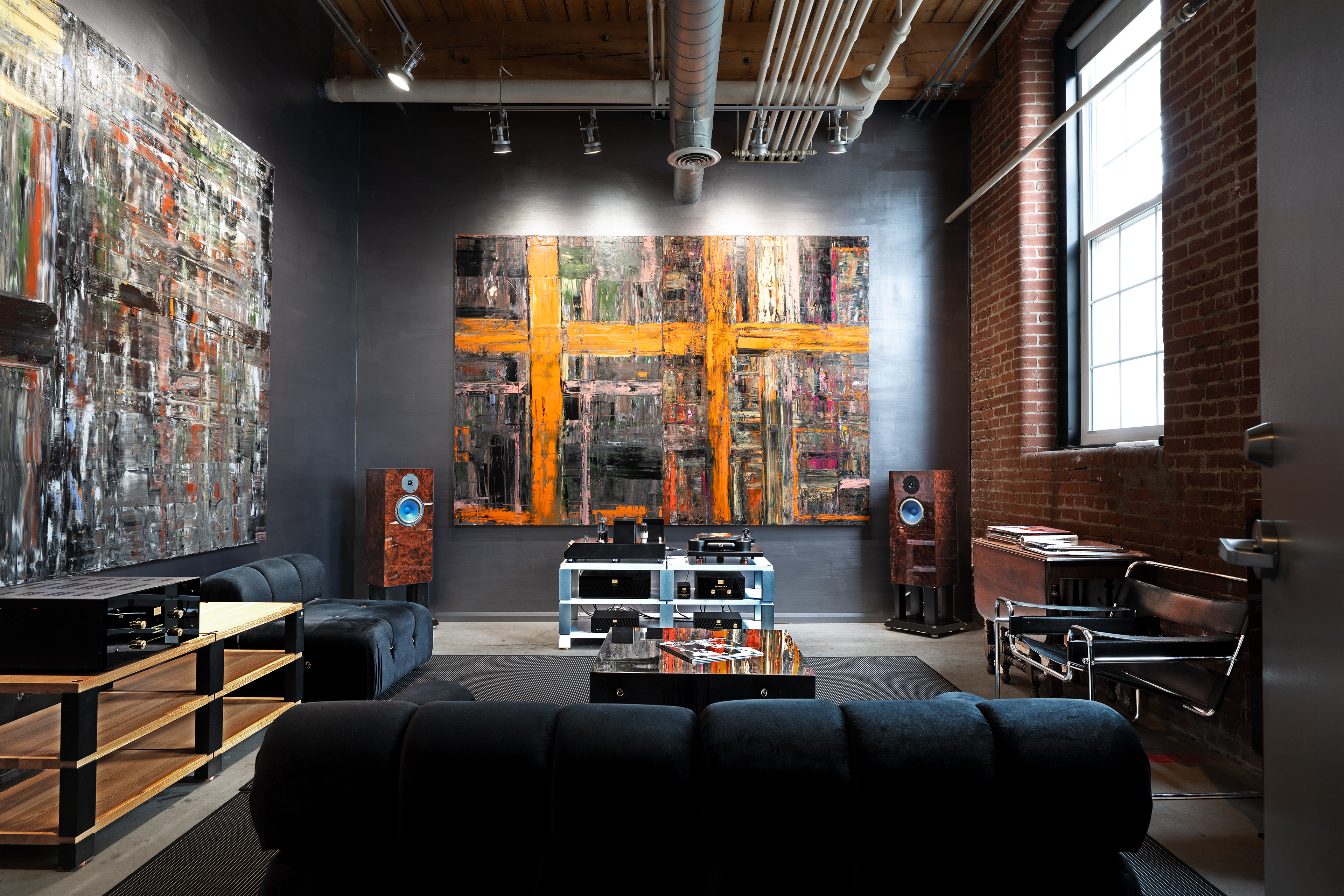 The Audio Autobahn floor — Burmester monitors flanking an orange-cross painting, leather LC1 chair on brick.
